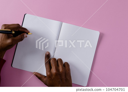 Hands of biracial man holding pen and writing in notebook with copy space on pink background Hands of biracial man holding pen and writing in notebook with copy space on pink background 104704701