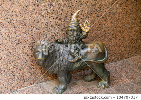 Dwarf riding a lion sleeping cap sitting on a lion and waving hello. Small bronze figures of gnomes on the streets of Wroclaw. Europe Miniature bronze figure of a gnome. Symbol of Wroclaw 104707017