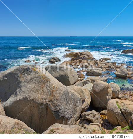 Beautiful seascape beach waves splashing against boulders or big stones in aqua sea water. A summer nature location in South Africa. Rocks in the ocean under a blue sky background with copy space. Beautiful seascape beach waves splashing against boulders or big stones in aqua sea water. A summer nature location in South Africa. Rocks in the ocean under a blue sky background with copy space. 104708584
