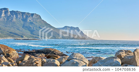 Quiet and calm ocean with rocky shore and boulders. Scenic seascape of the Twelve Apostles mountain under a blue sky in Cape Town. A beautiful holiday destination for travel and tourism Quiet and calm ocean with rocky shore and boulders. Scenic seascape of the Twelve Apostles mountain under a blue sky in Cape Town. A beautiful holiday destination for travel and tourism 104708636