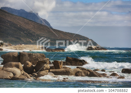 Ocean view of waves crashing against boulders or rocks in the sea in Camps Bay, Cape Town in South Africa. Relaxed and calm landscape view of the sea and mountains against a cloudy overcast sky 104708882