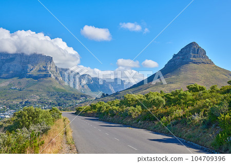 Panoramic and scenic view of an empty road in the countryside with mountains and greenery in summer. Beautiful landscape view of a quiet street surrounded by natural landmarks against a blue sky 104709396