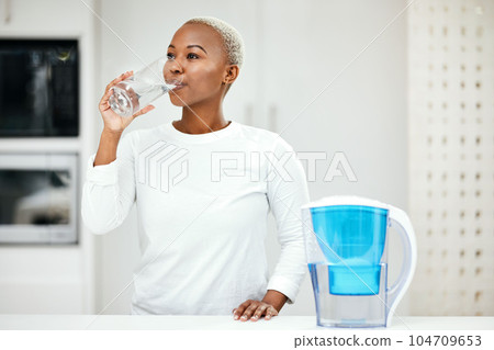 Woman, filter and drinking water for health at home for a healthy lifestyle, diet and nutrition. Young african person with a glass from a container or pitcher in a kitchen for detox and wellness Woman, filter and drinking water for health at home for a healthy lifestyle, diet and nutrition. Young african person with a glass from a container or pitcher in a kitchen for detox and wellness 104709653