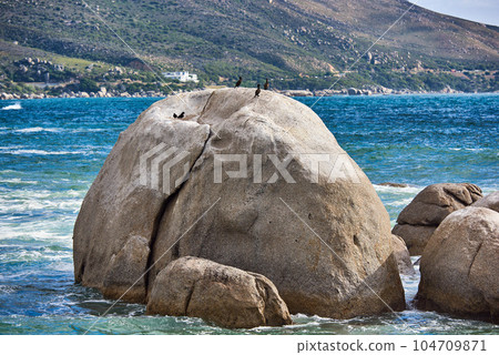 Big rocks in the blue ocean with mountains in the background. Stunning nature landscape or seascape on a summer day. Boulders or large natural stones in aqua sea water with beautiful rough textures 104709871