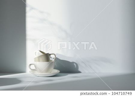 A room with various objects such as warm sunlight, shadows of grass leaves, a vase on a table, and cups 104710749