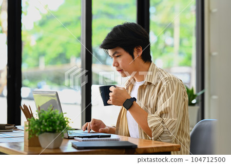 Asian businessman wearing smart casual wear drinking coffee and using laptop at office 104712500