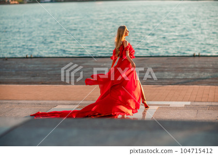 Sunrise red dress. A woman in a long red dress against the backdrop of sunrise, bright golden light of the sun's rays. The concept of femininity, harmony. 104715412
