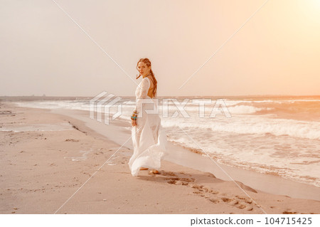 woman sea white dress. Model in boho style in a white long dress and silver jewelry on the beach. Her hair is braided, and there are many bracelets on her arms. 104715425