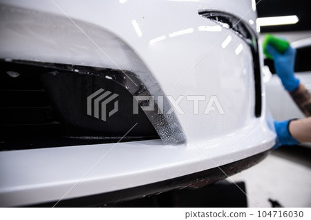 Car detailing studio employee, a wrapper, is protecting the bumper of a white car with a colorless protective PPF film. 104716030