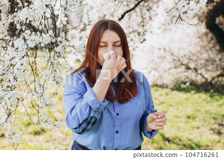Sneezing young redhead woman with nose wiper among blooming trees in park. Portrait of sick women sneezes in white tissue, suffers from rhinitis and running nose. Symptoms of cold or allergy. Sneezing young redhead woman with nose wiper among blooming trees in park. Portrait of sick women sneezes in white tissue, suffers from rhinitis and running nose. Symptoms of cold or allergy. 104716261