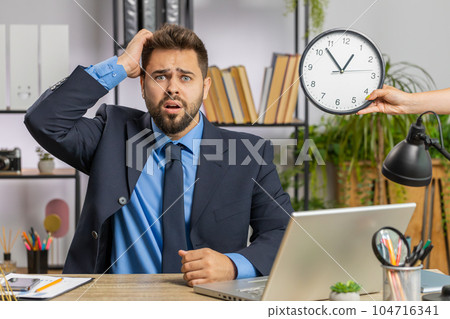 Young businessman with anxiety checking time on clock, running late to work being in delay deadline Young businessman with anxiety checking time on clock, running late to work being in delay deadline 104716341