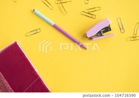Stationery is on a yellow background. A pen and a stapler are lying next to a pink notebook 104716390