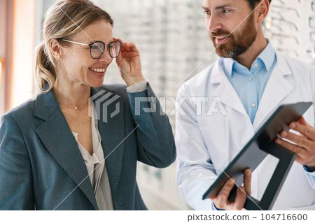 Close up of optometrist helping woman to choose glasses in optical store showing to her mirror Close up of optometrist helping woman to choose glasses in optical store showing to her mirror 104716600