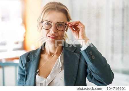 Focused Middle-aged woman in jacket trying to choose glasses in an optics store Focused Middle-aged woman in jacket trying to choose glasses in an optics store 104716601