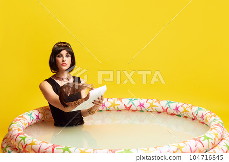 Serious young woman in image of Cleopatra sitting in swimming pool and writing letter against yellow studio background. 104716845