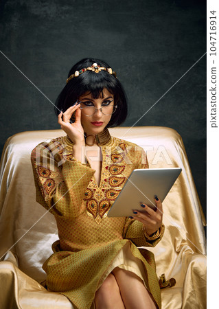 Portrait of young beautiful woman, queen, Cleopatra in golden dress sitting with tablet against dark vintage background. Business lady 104716914