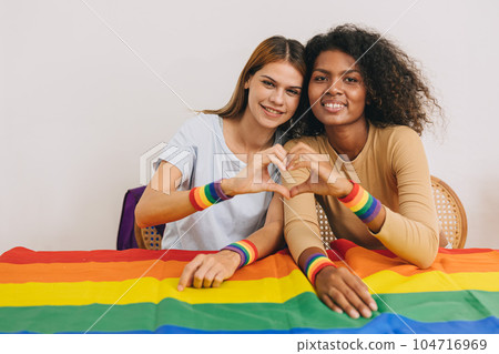 Portrait LGBT woman hand heart love sign mix race living together with rainbow flag looking camera 104716969