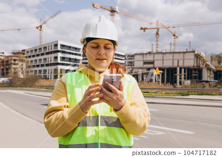 Young Architect woman in white hardhat and safety vest with digital tablet using smartphone outdoors. Female engineer. Logistics and construction worker near modern building. Engineering application Young Architect woman in white hardhat and safety vest with digital tablet using smartphone outdoors. Female engineer. Logistics and construction worker near modern building. Engineering application 104717182