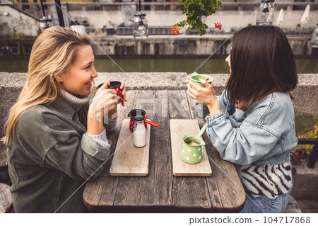 Two young women blonde and brunette sitting at a cafe by the river having coffee and a talk Two young women blonde and brunette sitting at a cafe by the river having coffee and a talk 104717868
