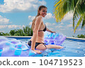 A young woman is relaxing by the pool. in the background a garden and palm trees. 104719523