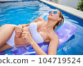 A young woman is relaxing by the pool. in the background a garden and palm trees. 104719524