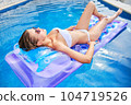 A young woman is relaxing by the pool. in the background a garden and palm trees. 104719526