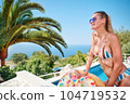 A young woman is relaxing by the pool. in the background a garden and palm trees. 104719532