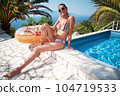 A young woman is relaxing by the pool. in the background a garden and palm trees. 104719533