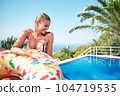 A young woman is relaxing by the pool. in the background a garden and palm trees. 104719535