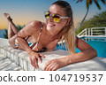 A young woman is relaxing by the pool. in the background a garden and palm trees. 104719537