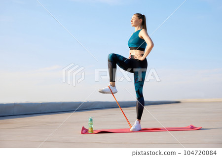 Leg exercises. Young sportive woman in stylish sportswear training fitn fitness elastic bands outdoors on warm sunny day 104720084