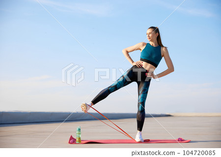 Leg exercises. Young sportive woman in stylish sportswear training fitn fitness elastic bands outdoors on warm sunny day 104720085