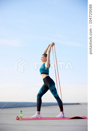 Feeling positive. Young sportive woman in stylish sportswear training fitn fitness elastic bands outdoors on warm sunny day 104720086