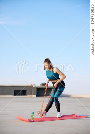 Strong hands. Young sportive woman in stylish sportswear training fitn fitness elastic bands outdoors on warm sunny day 104720089