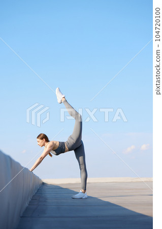 Flexibility and strength. Youn sportive woman in stylish sportswear training, doing stretching exercises outdoors on sunny day 104720100