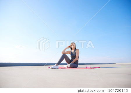 Young woman with slim, fit body sitting on fitness mantra in comfortable sportswear in sunny morning. Outdoor training 104720143