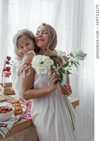A little blonde girl with her mom on a kitchen countertop decorated with peonies. The concept of the relationship between mother and daughter. Spring atmosphere. A little blonde girl with her mom on a kitchen countertop decorated with peonies. The concept of the relationship between mother and daughter. Spring atmosphere. 104723273