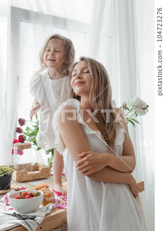 A little blonde girl with her mom on a kitchen countertop decorated with peonies. The concept of the relationship between mother and daughter. Spring atmosphere. A little blonde girl with her mom on a kitchen countertop decorated with peonies. The concept of the relationship between mother and daughter. Spring atmosphere. 104723276
