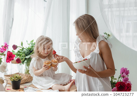 A little blonde girl with her mom on a kitchen countertop decorated with peonies. The concept of the relationship between mother and daughter. Spring atmosphere. A little blonde girl with her mom on a kitchen countertop decorated with peonies. The concept of the relationship between mother and daughter. Spring atmosphere. 104723277