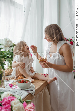 A little blonde girl with her mom on a kitchen countertop decorated with peonies. The concept of the relationship between mother and daughter. Spring atmosphere. 104723280