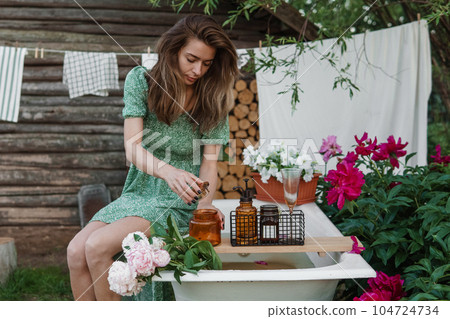 A woman is sitting on a cast-iron bathtub in the courtyard of a country house next to a bush of flowering peonies. The concept of summer, country life, a bathroom on the street in a blooming garden in 104724734