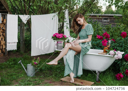 A woman is sitting on a cast-iron bathtub in the courtyard of a country house next to a bush of flowering peonies. The concept of summer, country life, a bathroom on the street in a blooming garden in A woman is sitting on a cast-iron bathtub in the courtyard of a country house next to a bush of flowering peonies. The concept of summer, country life, a bathroom on the street in a blooming garden in 104724736