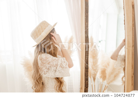 A beautiful teenage girl with long hair measures a straw hat in front of a mirror. Self-admiration of a blonde. 104724877