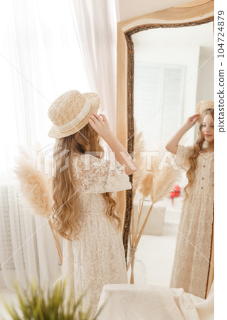 A beautiful teenage girl with long hair measures a straw hat in front of a mirror. Self-admiration of a blonde. 104724879