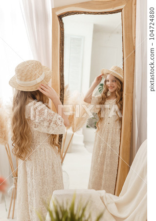 A beautiful teenage girl with long hair measures a straw hat in front of a mirror. Self-admiration of a blonde. A beautiful teenage girl with long hair measures a straw hat in front of a mirror. Self-admiration of a blonde. 104724880