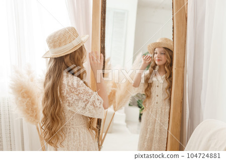 A beautiful teenage girl with long hair measures a straw hat in front of a mirror. Self-admiration of a blonde. A beautiful teenage girl with long hair measures a straw hat in front of a mirror. Self-admiration of a blonde. 104724881
