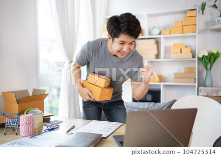 Happy man Celebrating Order from Internet Customer: New Business Style at Home, Online Selling, Young Entrepreneur's Success: Work-from-Home SME Owner Thrives in Online Sales, Packing Box. 104725314