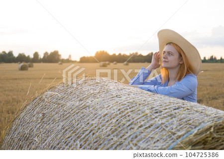 A red-haired woman in a hat and a blue dress walks in a field with haystacks. A red-haired woman in a hat and a blue dress walks in a field with haystacks. 104725386