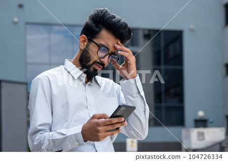 Young hispanic businessman outside office building, man in shirt reading sad bad news online using app on phone, depressed man disappointed unhappy with achievement results. 104726334