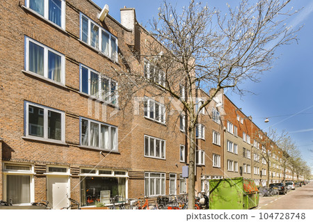 a tree in front of an apartment building with bicycles parked on the street next to it and people walking down the sidewalk 104728748